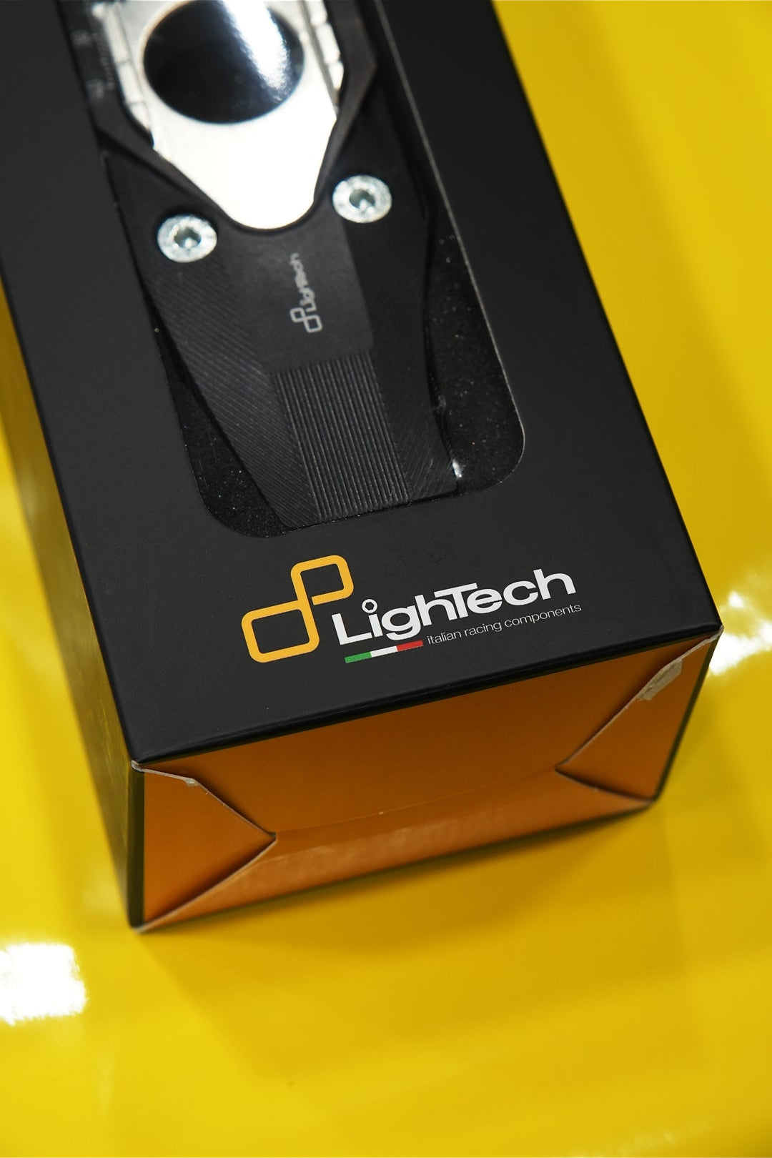 A close-up of a LightTech product in its black retail box, featuring a visible silver and black circular component through a clear window, set against a bright yellow background.